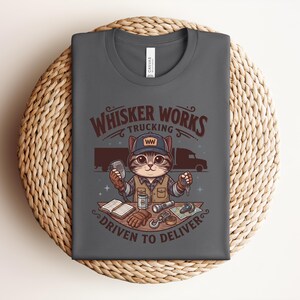 May include: Dark grey t-shirt with a cartoon cat trucker design. The text reads "Whisker Works Trucking" and "Driven to Deliver." The cat is depicted in a trucker hat, holding a CB radio and keys. The design includes a lorry and trucking-related accessories.