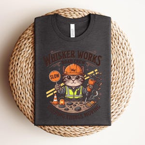 May include: Dark grey t-shirt with a cartoon cat in a construction worker outfit, holding a "SLOW" sign and a jackhammer. The shirt has the text "Whisker Works Road Crew" and "Keeping Things Moving".