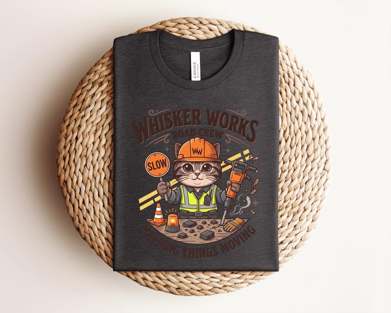May include: Dark grey t-shirt with a cartoon cat in a construction worker outfit, holding a "SLOW" sign and a jackhammer. The shirt has the text "Whisker Works Road Crew" and "Keeping Things Moving".