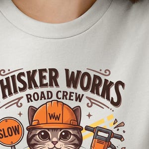 May include: A light grey t-shirt with a cartoon cat wearing an orange hard hat. The text "Whisker Works Road Crew" is above the cat, and a "SLOW" sign is to the left. A jackhammer is also visible.