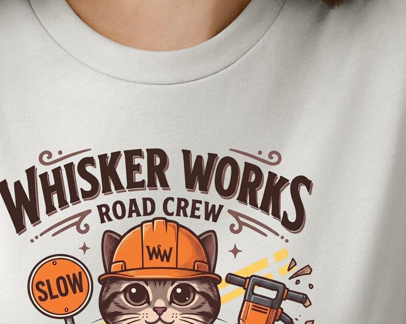 May include: A light grey t-shirt with a cartoon cat wearing an orange hard hat. The text "Whisker Works Road Crew" is above the cat, and a "SLOW" sign is to the left. A jackhammer is also visible.