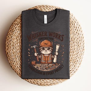 Puede incluir: Camiseta gris oscuro con un gato de dibujos animados con un atuendo de trabajador de la construcci&oacute;n y un casco. La camiseta tiene el texto "Whisker Works Construction" y "Purrcision in Every Project". El gato sostiene un martillo y un plano.