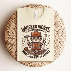 Puede incluir: Camiseta color crema con un gato de dibujos animados con un atuendo de trabajador de la construcci&oacute;n. El gato sostiene un martillo y un plano, con el texto "Whisker Works Construction" y "Purrcision in Every Project". La camiseta est&aacute; doblada sobre una estera tejida.