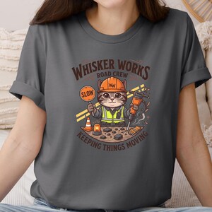 May include: Dark grey t-shirt with a cartoon cat dressed as a construction worker. The cat holds a "SLOW" sign and a jackhammer. The shirt features the text "Whisker Works Road Crew Keeping Things Moving."
