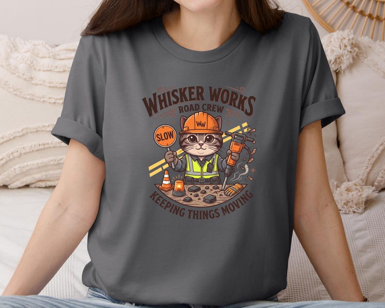 May include: Dark grey t-shirt with a cartoon cat dressed as a construction worker. The cat holds a "SLOW" sign and a jackhammer. The shirt features the text "Whisker Works Road Crew Keeping Things Moving."