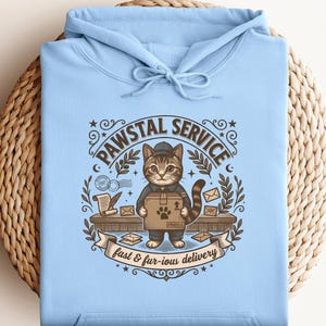 Op de afbeelding: Een lichtblauwe hoodie met een afbeelding van een cyperse kat verkleed als bezorger die een doos vasthoudt. De tekst luidt PAWSTAL SERVICE en fast & fur-ious delivery. Het ontwerp bevat enveloppen, een perkamentrol en decoratieve krullen.