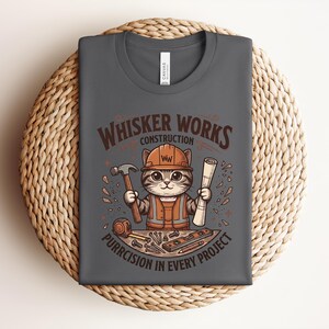 Puede incluir: Camiseta gris oscuro con un gato de dibujos animados con un atuendo de trabajador de la construcci&oacute;n, sosteniendo un martillo y planos. La camiseta dice "Whisker Works Construction" y "Purrcision in Every Project."