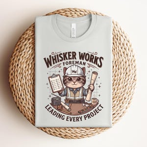 May include: Light gray t-shirt featuring a cartoon cat foreman wearing a hard hat and holding blueprints and a clipboard. The shirt has the text "Whisker Works Foreman Leading Every Project" in brown and white.