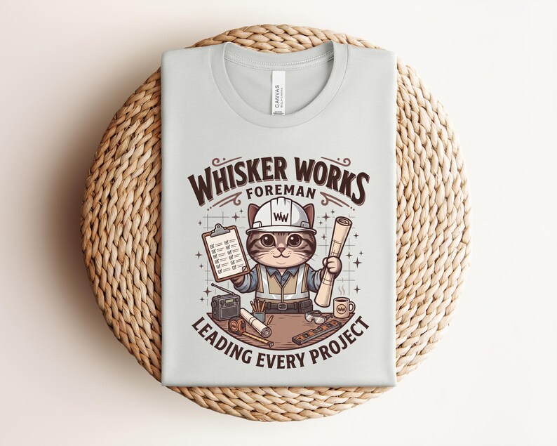 May include: Light gray t-shirt featuring a cartoon cat foreman wearing a hard hat and holding blueprints and a clipboard. The shirt has the text "Whisker Works Foreman Leading Every Project" in brown and white.