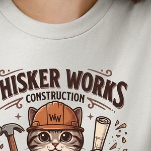 Puede incluir: Camiseta gris claro con un gato de dibujos animados con un casco de construcci&oacute;n. El gato sostiene un martillo y planos, con el texto "Whisker Works Construction" arriba. El dise&ntilde;o incluye detalles en marr&oacute;n y naranja.