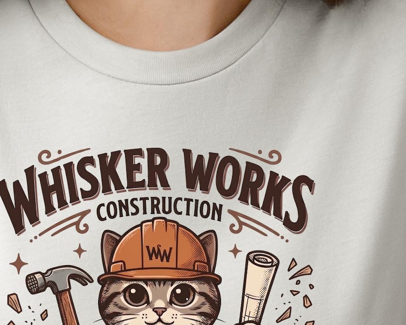 Puede incluir: Camiseta gris claro con un gato de dibujos animados con un casco de construcci&oacute;n. El gato sostiene un martillo y planos, con el texto "Whisker Works Construction" arriba. El dise&ntilde;o incluye detalles en marr&oacute;n y naranja.