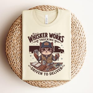 May include: Cream-coloured t-shirt with a cartoon cat trucker design. The text reads "Whisker Works Trucking" and "Driven to Deliver." The cat wears a trucker hat and holds a microphone and keys. A semi-truck is in the background.