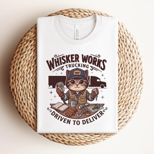 May include: White t-shirt with a cartoon cat trucker design. The text reads "Whisker Works Trucking" and "Driven to Deliver". The cat is wearing a cap and holding a CB radio and keys. The design includes a truck and trucking-related items.