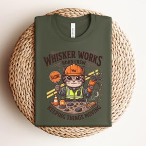 May include: Khaki green t-shirt with a cartoon cat dressed as a road worker. The cat holds a "SLOW" sign and a jackhammer. The shirt displays the text "Whisker Works Road Crew" and "Keeping Things Moving."