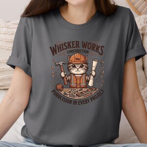 Puede incluir: Camiseta gris oscuro con un gato de dibujos animados con un atuendo de trabajador de la construcci&oacute;n, sosteniendo un martillo y planos. La camiseta tiene el texto "Whisker Works Construction" y "Purrcision in Every Project" en un dise&ntilde;o circular.