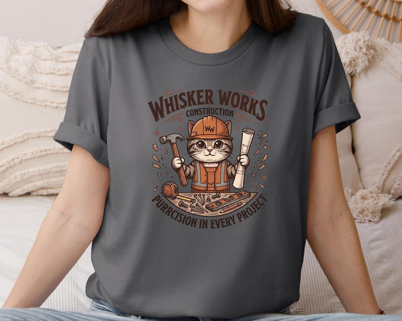 Puede incluir: Camiseta gris oscuro con un gato de dibujos animados con un atuendo de trabajador de la construcci&oacute;n, sosteniendo un martillo y planos. La camiseta tiene el texto "Whisker Works Construction" y "Purrcision in Every Project" en un dise&ntilde;o circular.