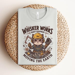 May include: A light gray t-shirt with a graphic of a cat wearing a construction hat and overalls. The text "Whisker Works Heavy Equipment Moving the Earth" is above and below the cat illustration. The shirt is folded on a woven, circular surface.
