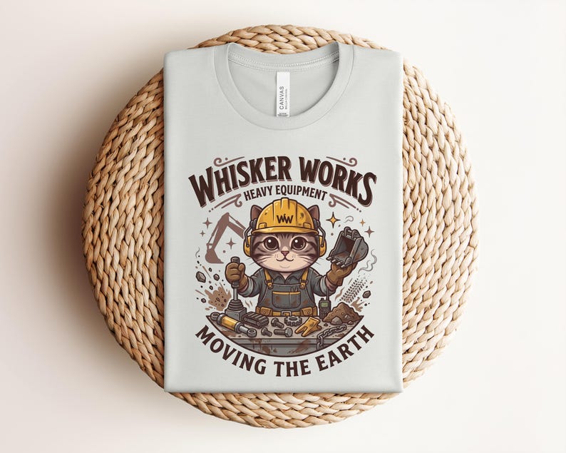 May include: A light gray t-shirt with a graphic of a cat wearing a construction hat and overalls. The text "Whisker Works Heavy Equipment Moving the Earth" is above and below the cat illustration. The shirt is folded on a woven, circular surface.