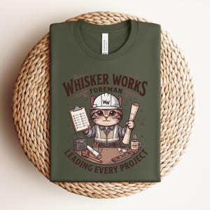 May include: Olive green t-shirt featuring a cartoon cat in a construction worker outfit. The cat is holding blueprints and a clipboard, with the text "Whisker Works Foreman Leading Every Project". The shirt is folded on a woven mat.