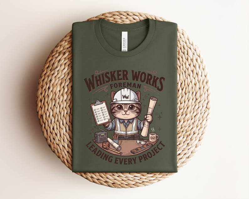 May include: Olive green t-shirt featuring a cartoon cat in a construction worker outfit. The cat is holding blueprints and a clipboard, with the text "Whisker Works Foreman Leading Every Project". The shirt is folded on a woven mat.