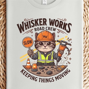 May include: A pale grey t-shirt with a cartoon cat wearing an orange hard hat and safety vest, holding a "SLOW" sign and a jackhammer. The text "WHISKER WORKS ROAD CREW KEEPING THINGS MOVING" is arched above and below the cat illustration.