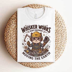 May include: White t-shirt featuring a cartoon cat wearing a construction hat and overalls. The text "Whisker Works Heavy Equipment Moving the Earth" is printed above and below the cat illustration. The shirt is folded on a woven, circular surface.