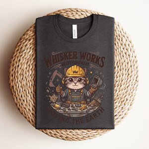 May include: Dark gray t-shirt featuring a cartoon cat wearing a yellow hard hat and overalls, holding tools. The shirt has the text "Whisker Works Heavy Equipment Moving the Earth" in brown and white.