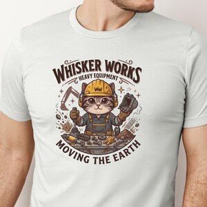 May include: White t-shirt featuring a cartoon cat wearing a yellow hard hat and overalls, holding construction tools. The shirt has the text "Whisker Works Heavy Equipment Moving the Earth" in brown lettering. The design includes an excavator and other construction elements.