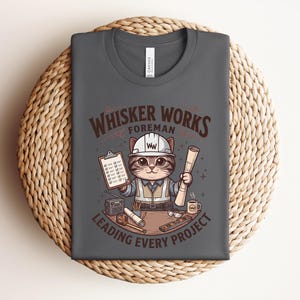 May include: Dark gray t-shirt featuring a cartoon cat in a construction worker outfit, holding blueprints and a clipboard. The shirt reads "Whisker Works Foreman Leading Every Project" in brown lettering. The shirt is folded on a woven, circular surface.