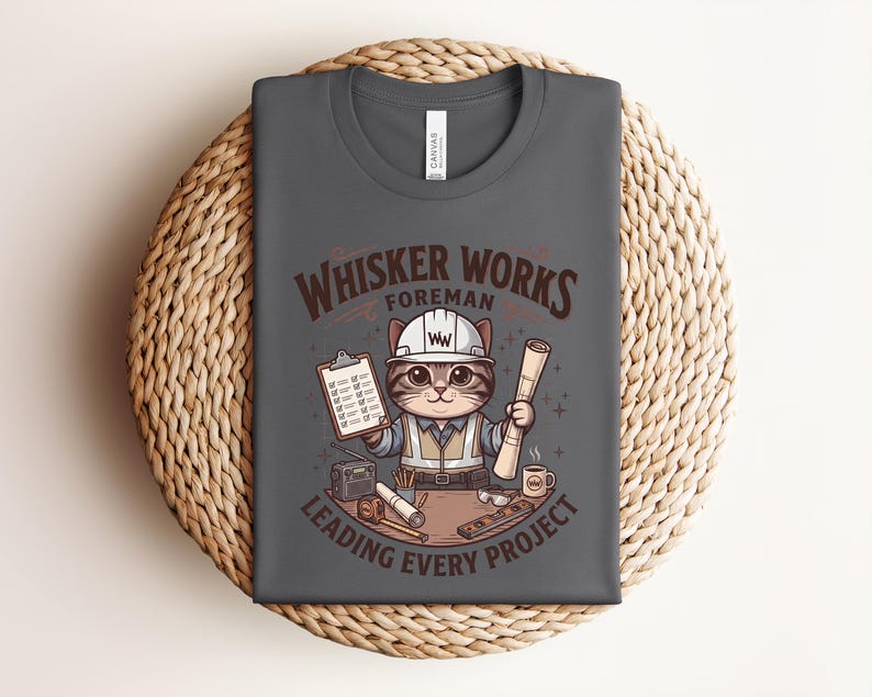 May include: Dark gray t-shirt featuring a cartoon cat in a construction worker outfit, holding blueprints and a clipboard. The shirt reads "Whisker Works Foreman Leading Every Project" in brown lettering. The shirt is folded on a woven, circular surface.