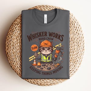 May include: Dark grey t-shirt with a cartoon cat in a construction worker outfit. The cat holds a "SLOW" sign and a jackhammer. Text on the shirt reads "WHISKER WORKS ROAD CREW KEEPING THINGS MOVING."