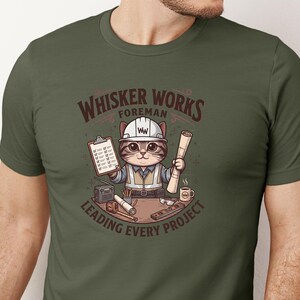 May include: Olive green t-shirt featuring a cartoon cat foreman wearing a hard hat and vest, holding blueprints and a clipboard. The text reads "Whisker Works Foreman Leading Every Project."