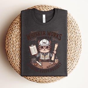 May include: Dark gray t-shirt featuring a cartoon cat foreman with a hard hat, holding blueprints and a clipboard. The text "Whisker Works Foreman" is above the cat, with "Finishing Every Project" below. The shirt is displayed on a woven surface.