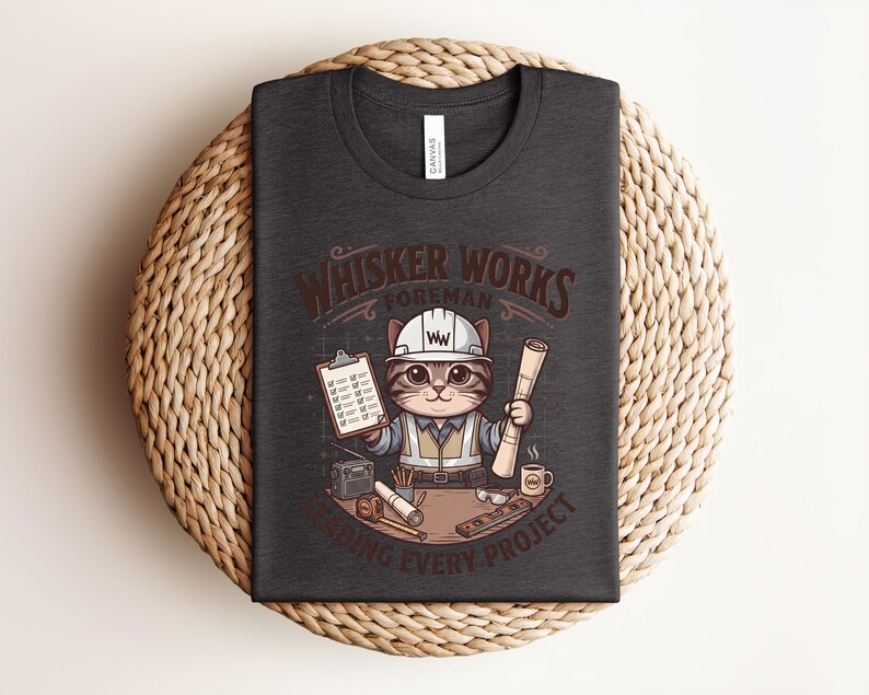 May include: Dark gray t-shirt featuring a cartoon cat foreman with a hard hat, holding blueprints and a clipboard. The text "Whisker Works Foreman" is above the cat, with "Finishing Every Project" below. The shirt is displayed on a woven surface.