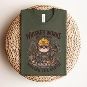 May include: Olive green t-shirt featuring a cartoon cat wearing a construction hat and overalls. The shirt has the text "Whisker Works Heavy Equipment Moving the Earth" in a brown, vintage-style font. The cat is surrounded by construction tools.