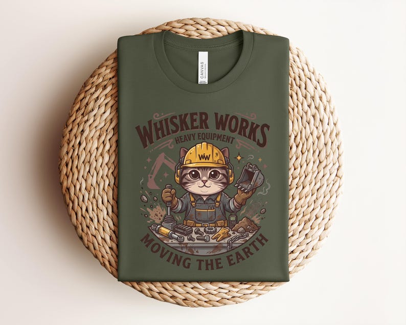 May include: Olive green t-shirt featuring a cartoon cat wearing a construction hat and overalls. The shirt has the text "Whisker Works Heavy Equipment Moving the Earth" in a brown, vintage-style font. The cat is surrounded by construction tools.