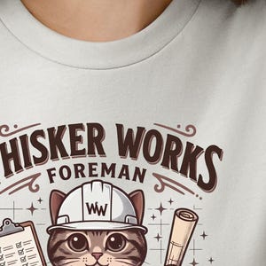 May include: A light grey t-shirt featuring a cartoon cat wearing a white hard hat with the letters "WW". The text above the cat reads "Whisker Works Foreman". The cat is holding a clipboard and a rolled-up blueprint.