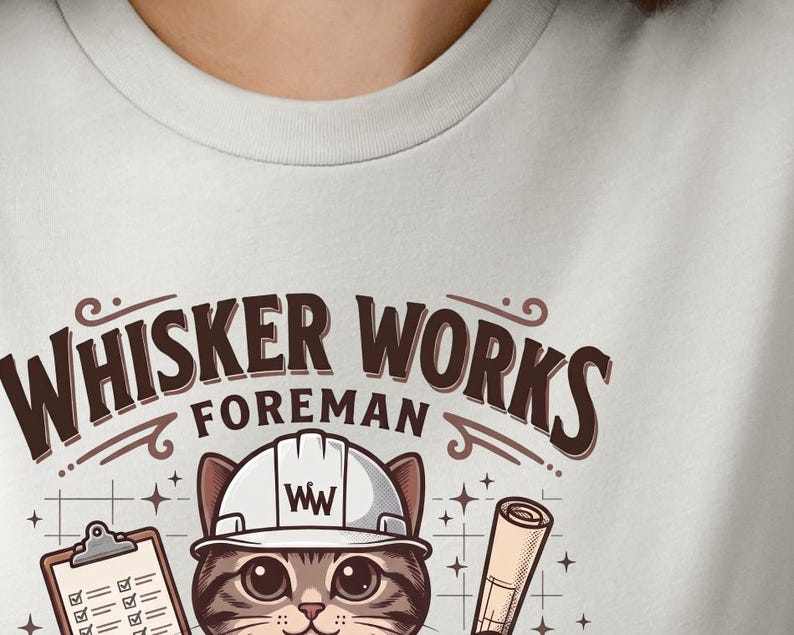 May include: A light grey t-shirt featuring a cartoon cat wearing a white hard hat with the letters "WW". The text above the cat reads "Whisker Works Foreman". The cat is holding a clipboard and a rolled-up blueprint.