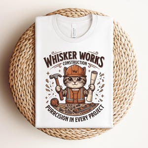 Puede incluir: Camiseta blanca con un gato de dibujos animados con casco y chaleco de construcci&oacute;n, sosteniendo un martillo y un plano. El texto dice "Whisker Works Construction" y "Purrcision in Every Project."