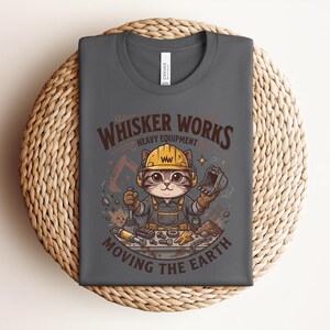 May include: Dark gray t-shirt featuring a cartoon cat wearing a construction helmet and overalls. The shirt has the text "Whisker Works Heavy Equipment Moving the Earth" in brown and orange. The cat is holding tools.