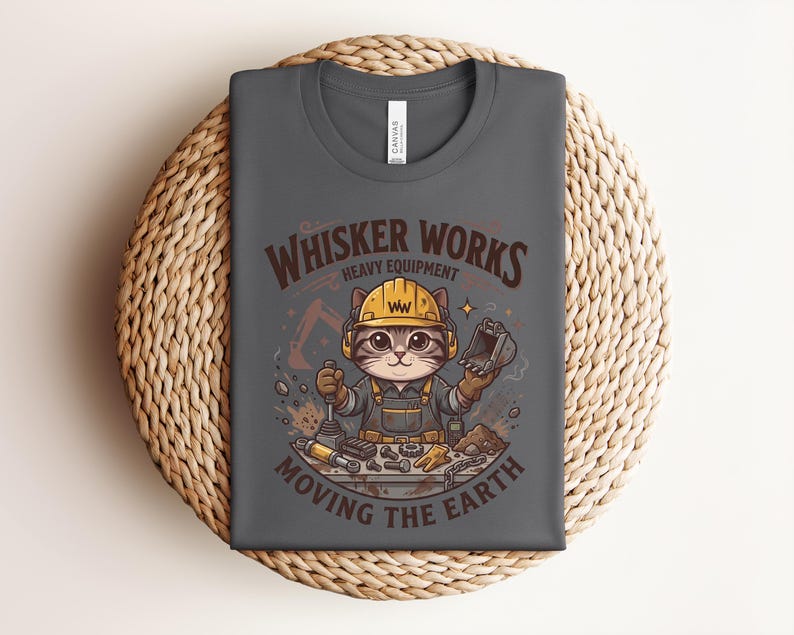 May include: Dark gray t-shirt featuring a cartoon cat wearing a construction helmet and overalls. The shirt has the text "Whisker Works Heavy Equipment Moving the Earth" in brown and orange. The cat is holding tools.