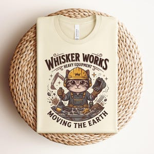 May include: Cream-colored t-shirt featuring a cartoon cat wearing a construction helmet and overalls. The text "Whisker Works Heavy Equipment Moving the Earth" is displayed above and below the cat illustration. The shirt is folded on a woven surface.