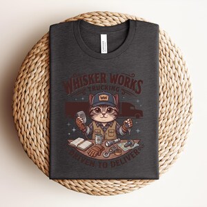 May include: Dark grey t-shirt featuring a cartoon cat trucker with a cap, microphone, and keys. The shirt displays the text "Whisker Works Trucking" and "Driven to Deliver". The design includes a truck and trucking-related items.