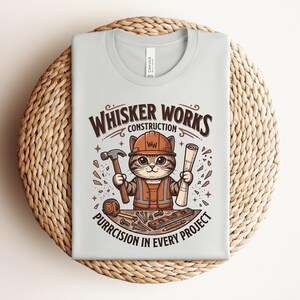 Puede incluir: Camiseta gris claro con un gato de dibujos animados con casco y chaleco de construcci&oacute;n, sosteniendo un martillo y un plano. El texto "Whisker Works Construction" y "Purrcision in Every Project" est&aacute; impreso en la camiseta.