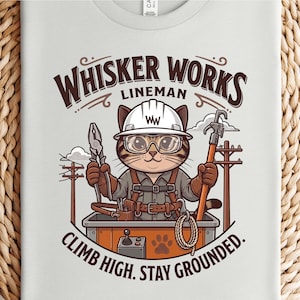 Puede incluir: Una camiseta de color claro con un gráfico de un gato vestido de liniero, con casco blanco y gafas de seguridad, sosteniendo herramientas. El texto dice "WHISKER WORKS LINEMAN" y "CLIMB HIGH. STAY GROUNDED." junto a postes eléctricos.