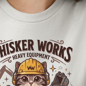 May include: A light gray t-shirt featuring a cartoon cat wearing a yellow construction helmet and headphones. The text "WHISKER WORKS HEAVY EQUIPMENT" is arched above the cat. The cat is surrounded by construction equipment and stars.