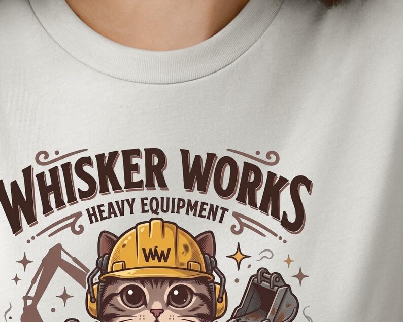 May include: A light gray t-shirt featuring a cartoon cat wearing a yellow construction helmet and headphones. The text "WHISKER WORKS HEAVY EQUIPMENT" is arched above the cat. The cat is surrounded by construction equipment and stars.