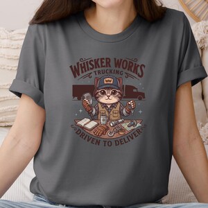 May include: Dark grey t-shirt with a cartoon cat in a trucker hat. The design includes the text "Whisker Works Trucking" and "Driven to Deliver." The cat is surrounded by trucking-related items, including a truck, keys, and a log book.