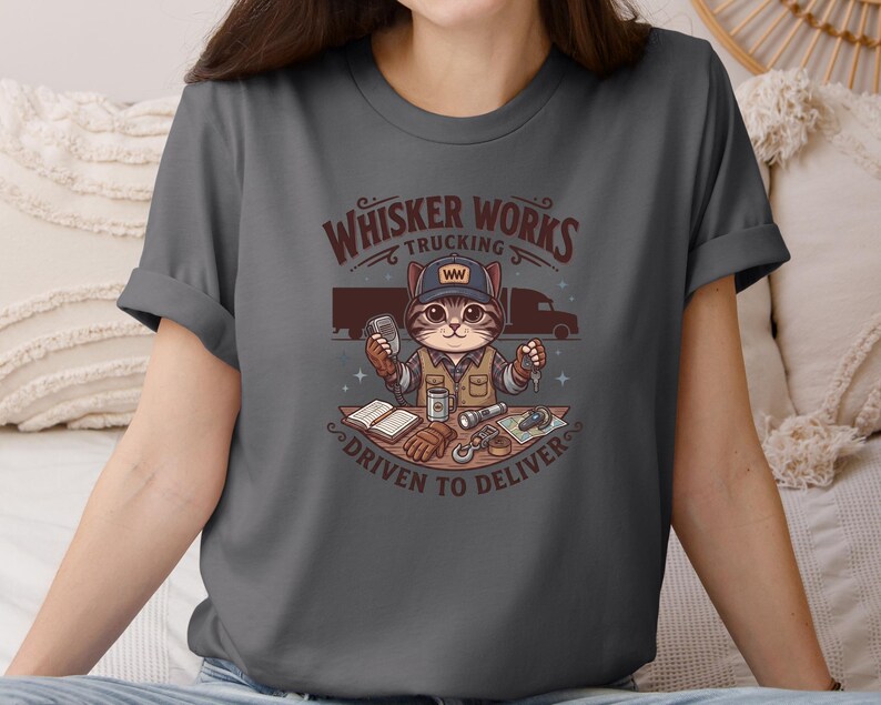 May include: Dark grey t-shirt with a cartoon cat in a trucker hat. The design includes the text "Whisker Works Trucking" and "Driven to Deliver." The cat is surrounded by trucking-related items, including a truck, keys, and a log book.