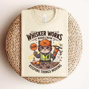 May include: A cream-coloured t-shirt with a cartoon cat dressed as a road worker. The cat holds a "SLOW" sign and a jackhammer. Text on the shirt reads "Whisker Works Road Crew" and "Keeping Things Moving."